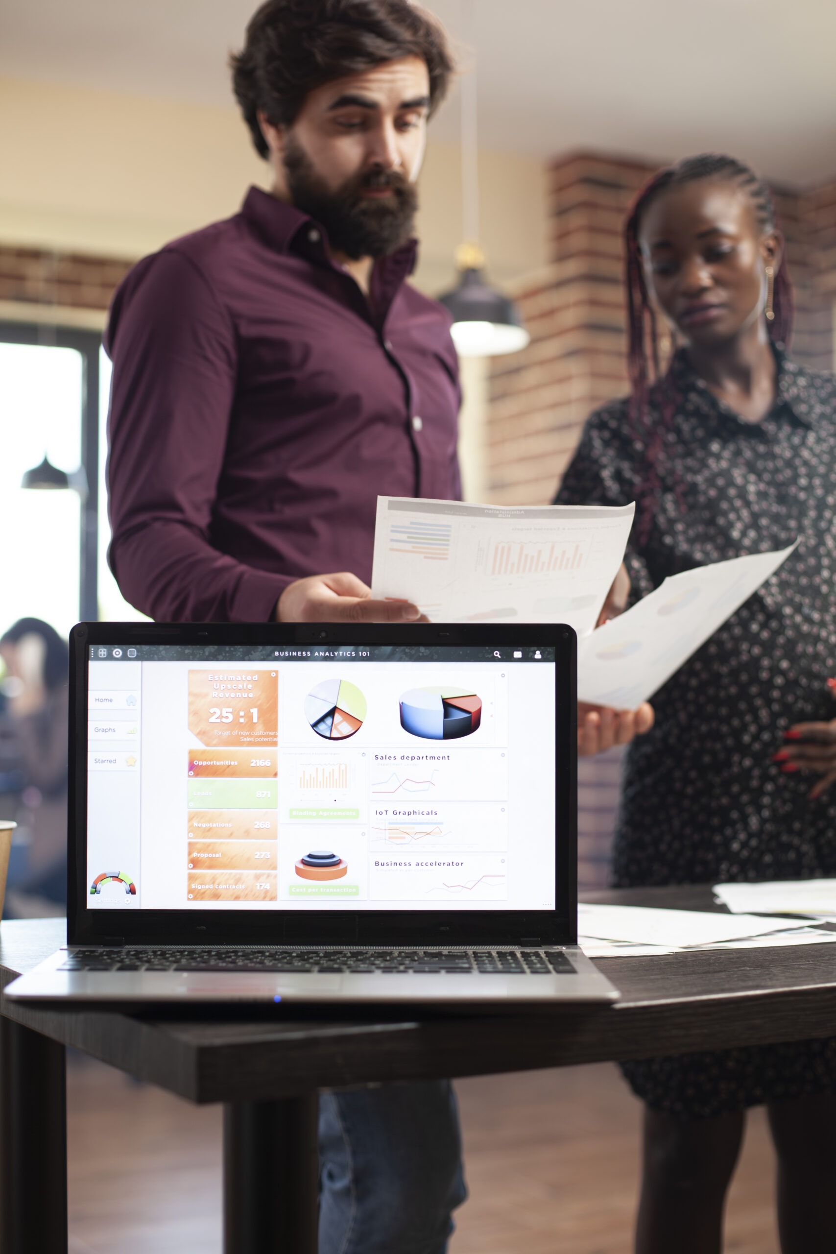 Pregnant lady and male colleague prepare market strategy as digital device shows data charts for research. Selective focus on laptop displaying financial analysis as diverse coworkers review documents